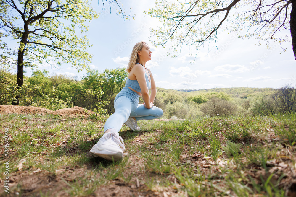 Naklejka premium The girl is doing a stretching squat on one leg. Beautiful blonde Caucasian woman in blue tight tracksuit. Blonde girl at an outdoor training session