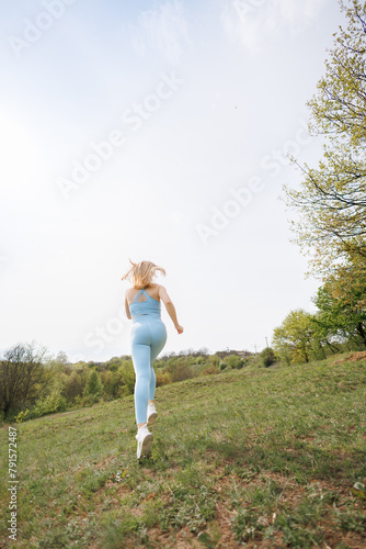 An athletic girl performs a jog in the park. Beautiful blonde Caucasian woman in blue tight tracksuit. Blonde girl at an outdoor training session