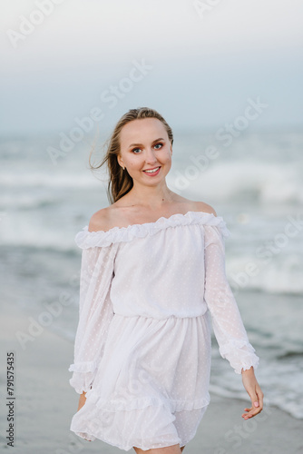 Happy woman go on seashore. Female stand on the beach ocean and enjoy summer day on vacation. Portrait of a beautiful girl in a white dress. Girl walks in water with big waves on sand sea in evening.