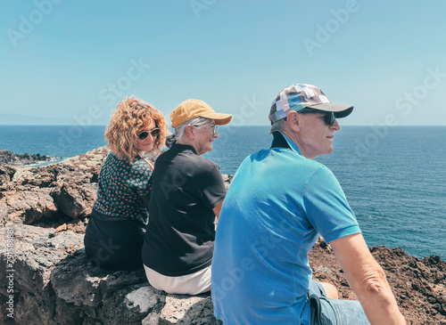 Back view of three friends in outdoor excursion at sea, sitting on the cliff looking at the horizon over water, enjoying freedom and vacation, healthy activity in retirement