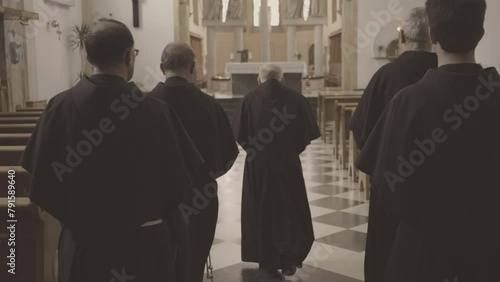 Christian priests walking along church pew rows aisle back view tracking shot. Monks waring robes moving to altar in temple. Christianity concept photo