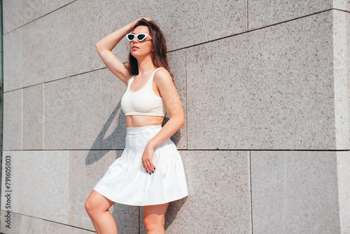 Young beautiful smiling hipster woman in trendy summer white tennis skirt and tank top clothes. Carefree woman posing in street at sunset. Positive model outdoors. Cheerful and happy, in sunglasses