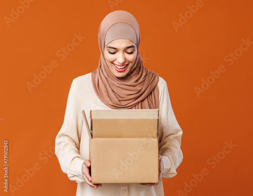 Beautiful overjoyed young smiling muslim woman in traditional religious hijab  holding an open cardboard box.