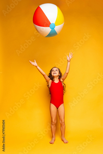 Happy little girl wearing red swimsuit and sunglasses playing with ball on yellow background. Summer vacation and childhood concept. Active lifestyle