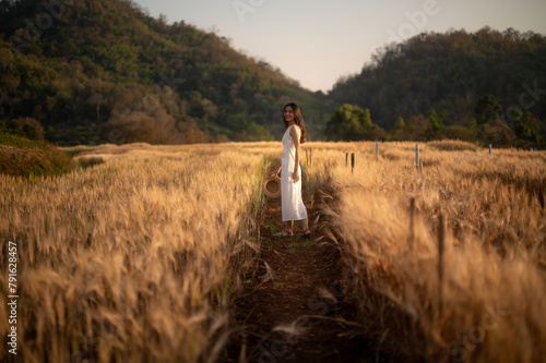 Fototapeta Naklejka Na Ścianę i Meble -  A young woman in a white dress and straw hat smiles while standing amidst a field of golden wheat, with a soft-focus mountain backdrop.