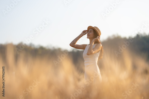 A young woman in a white dress and straw hat smiles while standing amidst a field of golden wheat, with a soft-focus mountain backdrop.