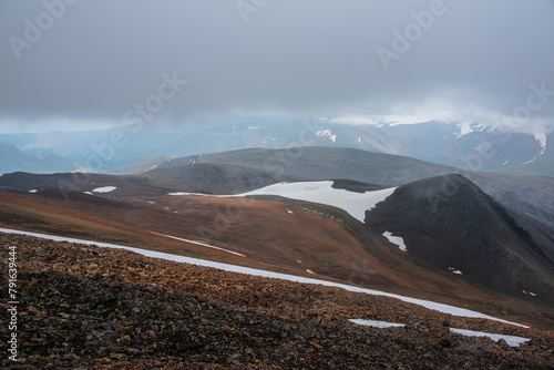 Dark overcast landscape with high pass, wide glacier on stony hill slope and snowy mountain range silhouette in far away in rainy low clouds. Large mountains with snow under gray sky in bad weather.