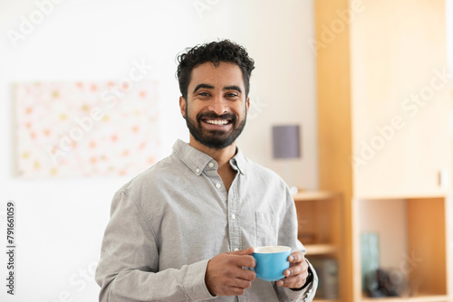 A smiling man with a beard holding a blue mug stands in a bright room with shelves photo