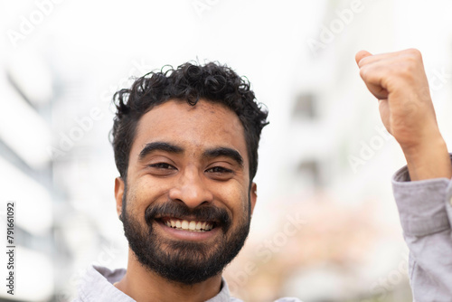 A smiling man with curly hair is raising his fist in a gesture of success or celebration against a blurred urban background. photo