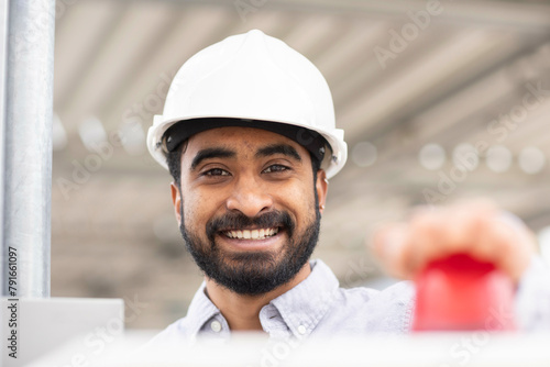 A smiling man in a white safety helmet is pressing a red button, with a steel structure in the background. photo