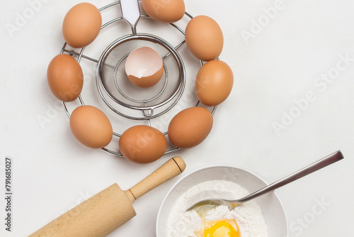 Brown eggs on metal stand. Egg yolk with flour and spoon in bowl. Milk in bowl and rolling pin on table