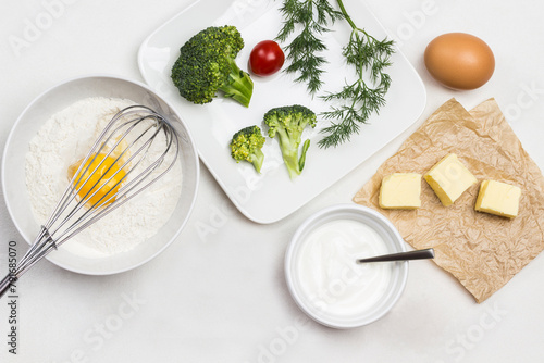 Ingredients for making dough. Broken egg with flour in bowl. Whisk on bowl. Butter on paper. Broccoli, tomatoes and dill in plate.