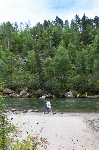 A tourist couple in love kisses on sandy beach of beautiful river against backdrop of green forest on vacation on sunny day. Summer trips and travels. River Ekhe-Ukhgun, Tunka, Buryatia, Baikal region photo