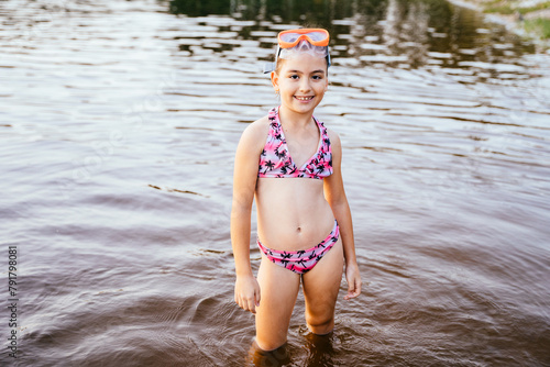 Happy preteen girl in swimsuit and mask in lake water standing knee-deep in water. Summertime children healthy vacation concept.