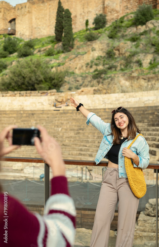 Two tourist friends in Malaga. One, with her back turned, photographs the other, who poses smiling and points to the monument with one arm.