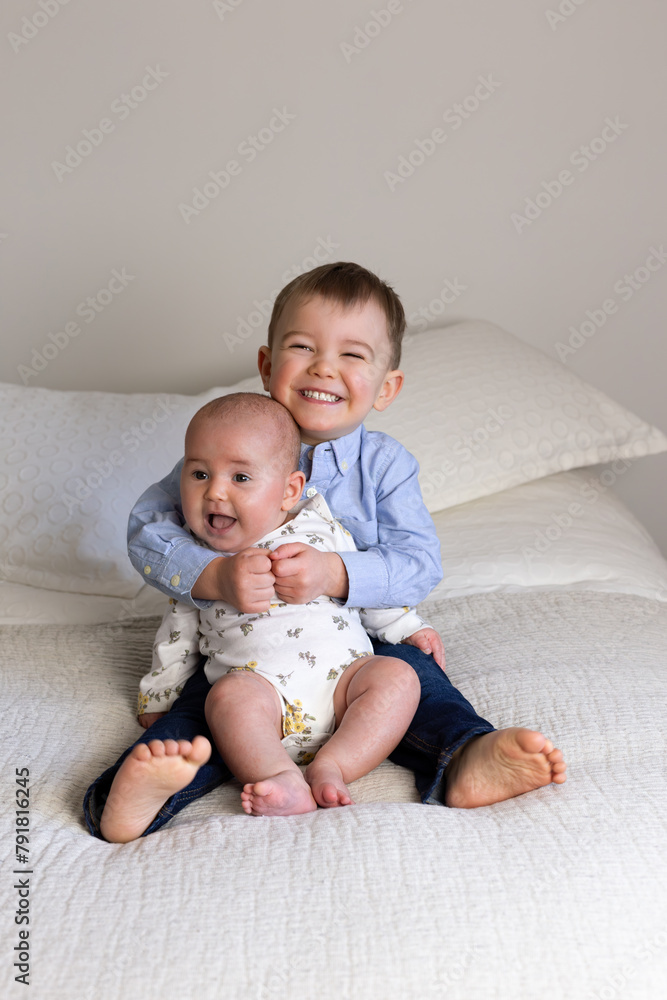 Vertical view of adorable smiling toddler boy sitting on bed holding his baby sister, Quebec City, Quebec, Canada Vertical view of adorable smiling toddler boy sitting on bed holding his baby sister, Quebec City, Quebec, Canada