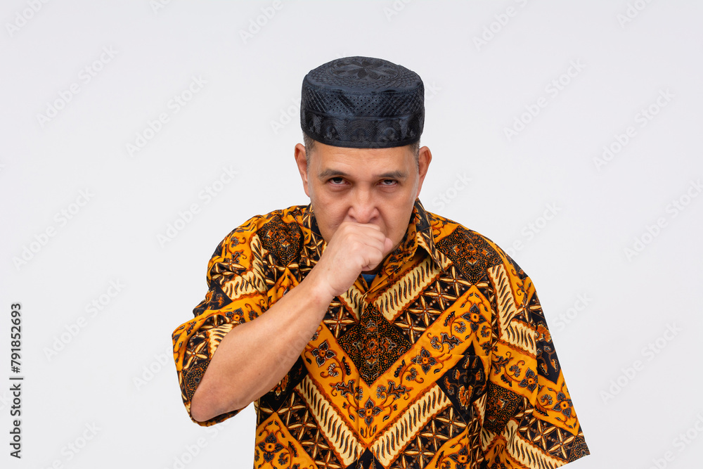 Fototapeta premium An Indonesian man in traditional batik shirt and kopiah hat looking angry while coughing, isolated on a white background.