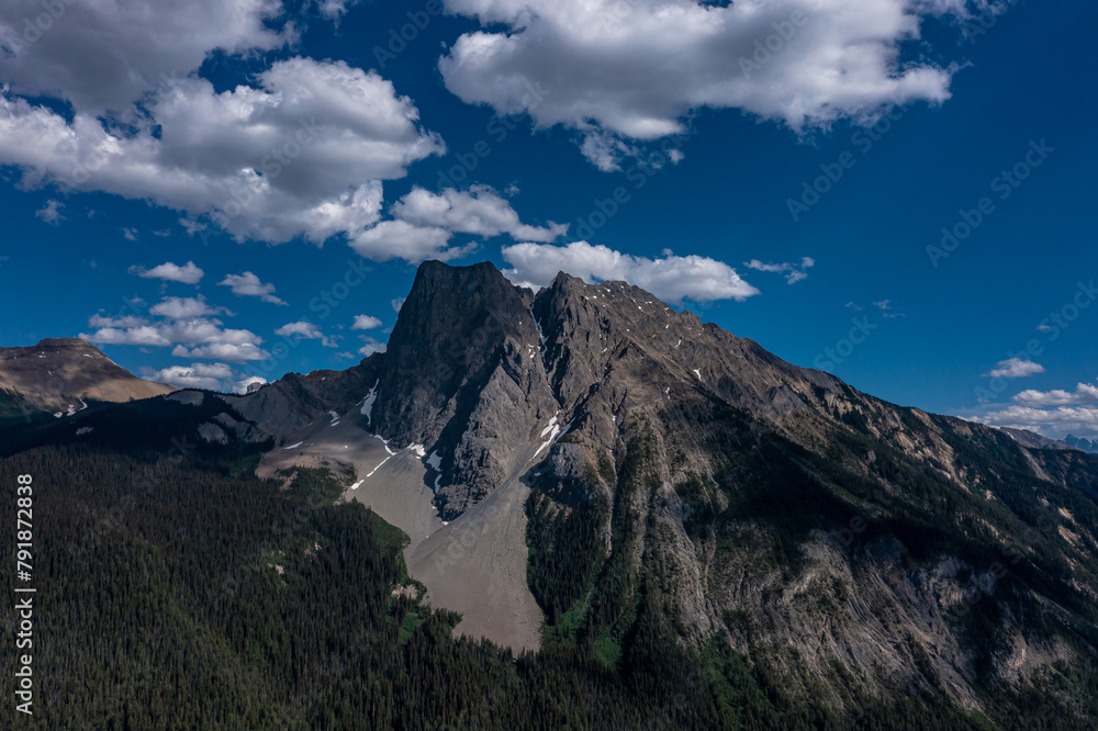 Drone view of mountains with high cliffs covered in forests under blue sky with white clouds. Canada
