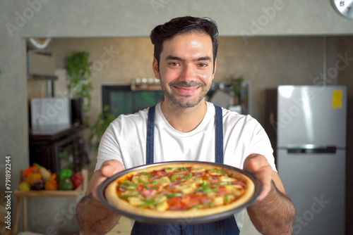 cheerful male chef presents a freshly baked pizza tray and smiles at the camera in the kitchen.