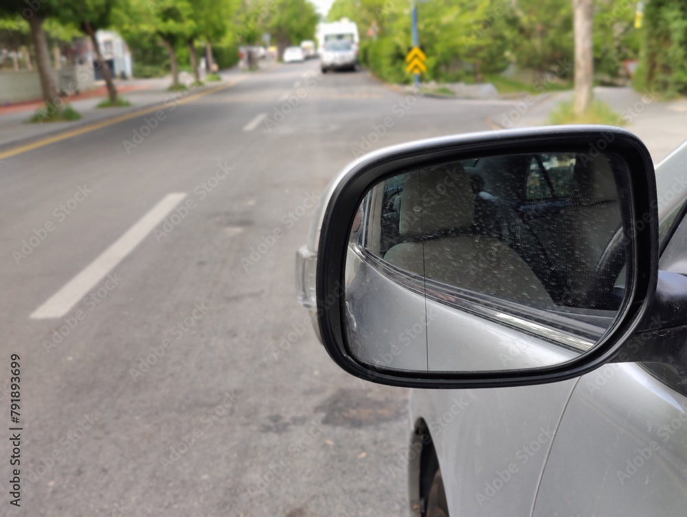 It shows a street scene seen from the perspective of a car's side mirror. The mirror reflects the interior of the car, including the side mirror, which is angled to show the road ahead.