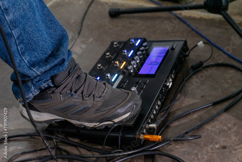 A foot steps on a guitar effects pedal, captured from the side. The shoe is a simple sneaker, and the pedal rests on a textured floor. The focus is on the action, with a sense of anticipation.