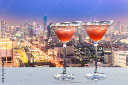 two red cocktails with ice cubes, blurred lights of Bangkok in the background