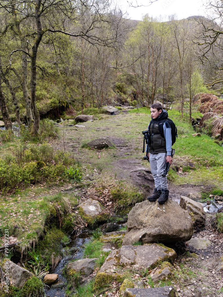 Young photographer standing on a large rock in a forest woodland scene with a slow moving moorland stream surrounded by trees Young photographer standing on a large rock in a forest woodland scene with a slow moving moorland stream surrounded by trees