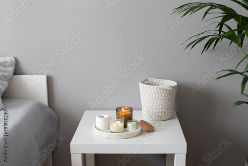 Home decor, stylish interior. Burning candles on marble tray standing on white badside table in bedroom.  Bed with grey blanket and pillows, wicker basket.