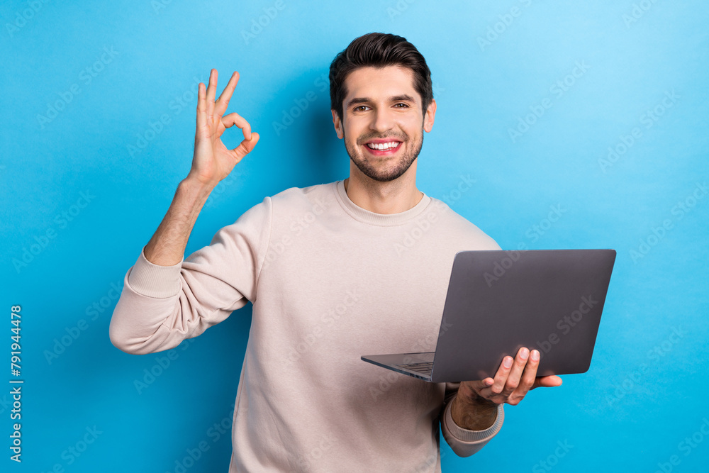 Photo of young handsome guy hold netbook show okey symbol wear beige sweatshirt isolated on blue color background Photo of young handsome guy hold netbook show okey symbol wear beige sweatshirt isolated on blue color background