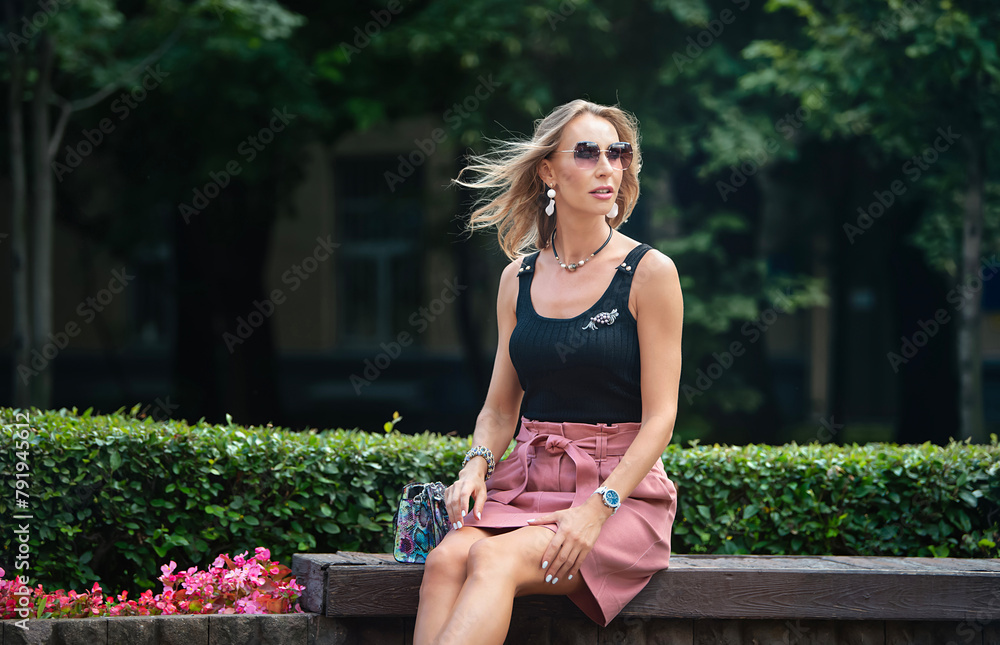 A stylish woman is waiting for a date on a bench in a city park. Woman in a black tight tank top with a deep neckline and a powder pink short denim skirt. A stylish woman is waiting for a date on a bench in a city park. Woman in a black tight tank top with a deep neckline and a powder pink short denim skirt.