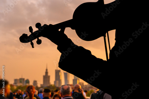 Sylwetka, grający na skrzypcach muzyk, zbierający pieniądze na tle Warszawskiego centrum o zachodzie słońca. Zdjęcie z mostu na centrum Warszawy © smoku7979