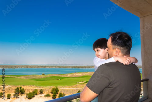 a man holds a little girl in his arms on a balcony against the backdrop of a golf course and the sea in the city of Abu Dhabi, a man and a child on vacation