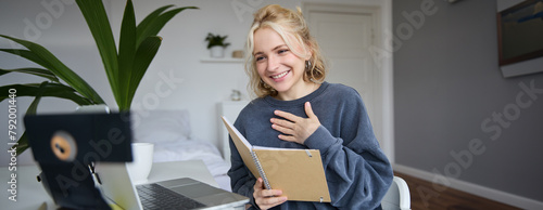 Portrait of young smiling blond woman, working from home, online chatting, using digital video camera, recording vlog, holding notebook, reading notes, explaining something