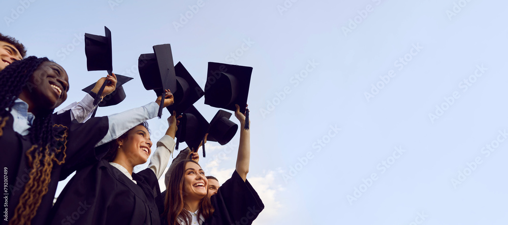 Group of happy multiethnic high school, college or university students having fun on graduation day and raising their graduate hats up to clear blue sky. Copy space banner background - obrazy, fototapety, plakaty 