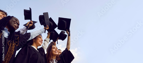 Group of happy multiethnic high school, college or university students having fun on graduation day and raising their graduate hats up to clear blue sky. Copy space banner background photo