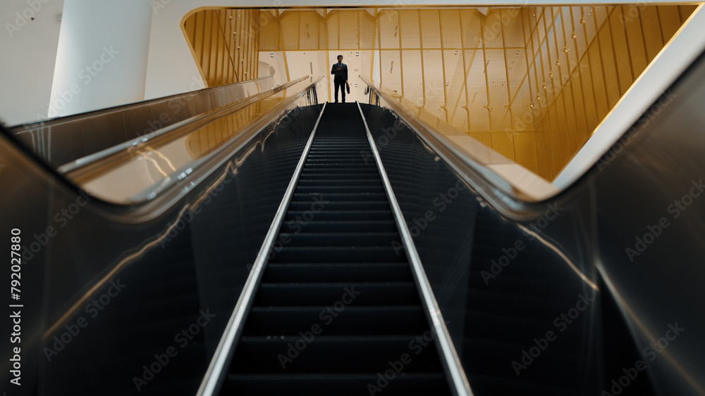 Fototapeta premium Professional business man using phone calling while using escalator. Skilled caucasian executive manager talking to business team to report and discuss marketing idea or plan. Front view. Exultant.