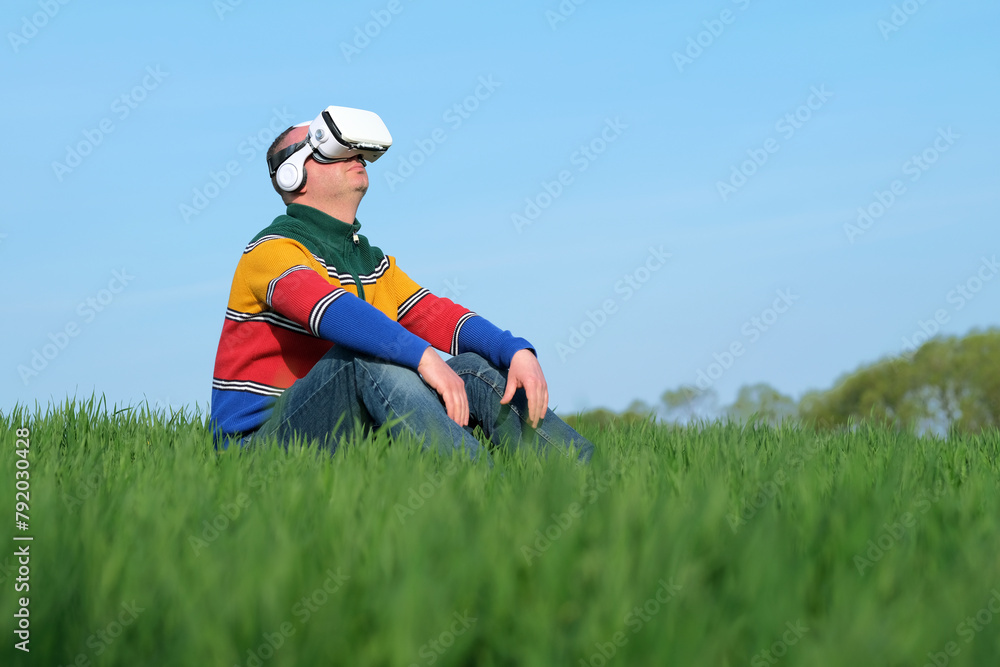 Man sitting on green grass in virtual reality glasses of the blue sky on the background, low viewing angle. Sustainable agriculture. outdoors Man sitting on green grass in virtual reality glasses of the blue sky on the background, low viewing angle. Sustainable agriculture. outdoors