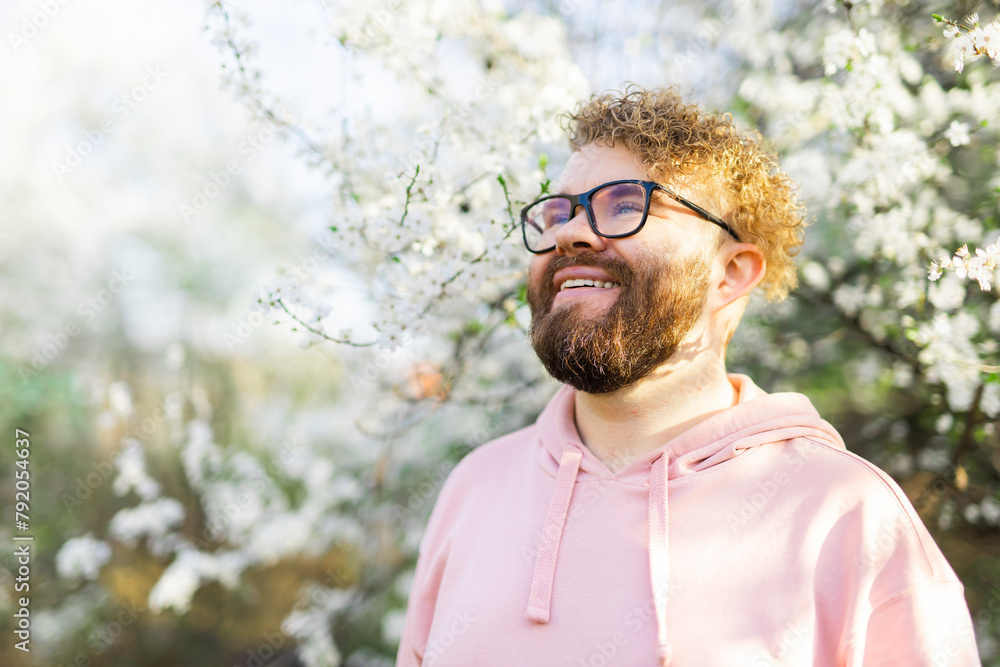 Male bearded man standing under branches with flowers of blooming almond or cherry tree in spring garden. Spring blossom. Copy space Male bearded man standing under branches with flowers of blooming almond or cherry tree in spring garden. Spring blossom. Copy space