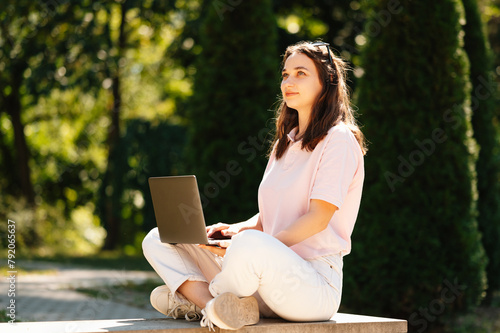 Cheerful female student is sitting and using her laptop in university campus or in park on a sunny day. photo