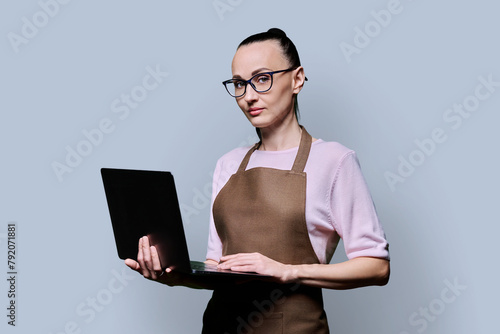 Portrait of 30s woman in apron holding laptop on grey background