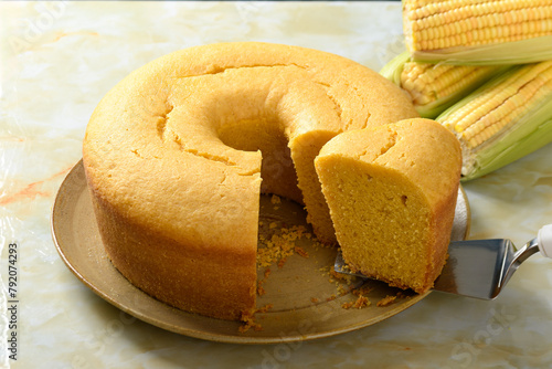  Corn cake on a plate on a yellow marble base, a slice being removed from the cake, ears of green corn on the table.