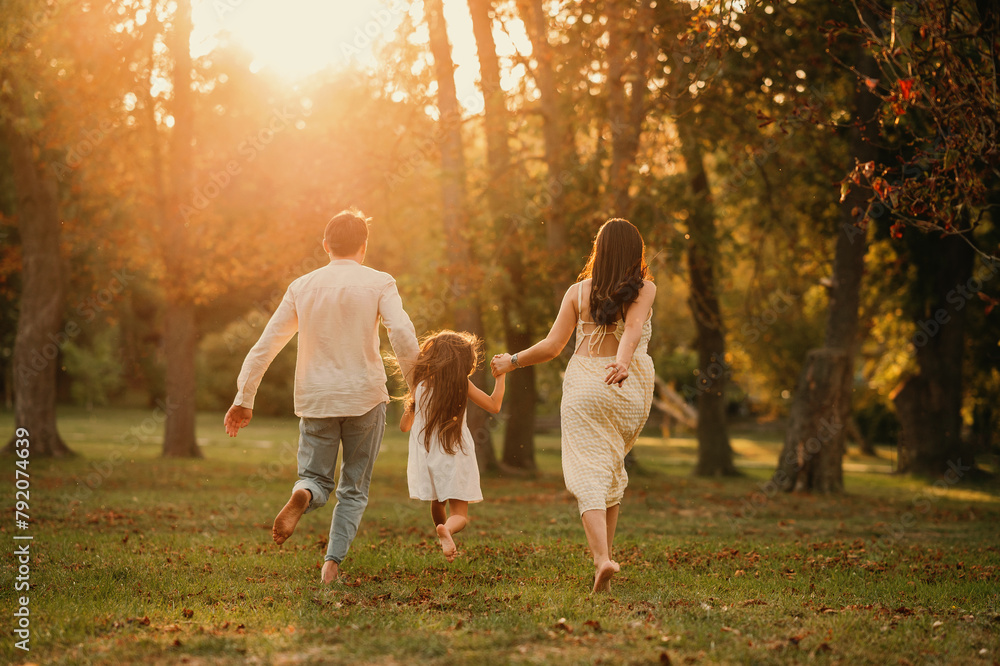 Young family of three are holding hands and running barefoot in a park together. Backview shot. Young family of three are holding hands and running barefoot in a park together. Backview shot.