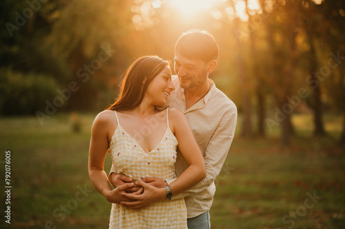 Young handsome man embraces his beautiful wife while laughing together in park at sunset.
