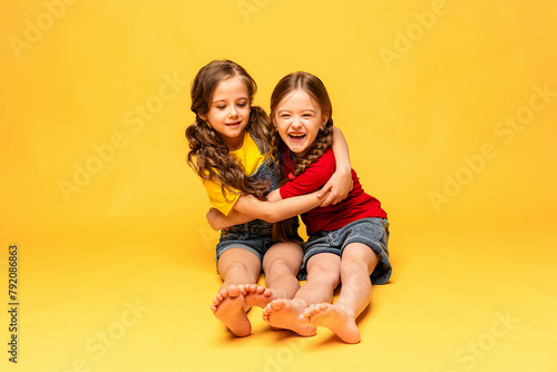 Two happy girls in bright summer clothes hugging and posing on a yellow background at studio. Summer children's fashion. Happy childhood. Advertising of children's products and sale