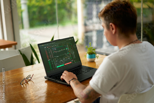 Focused crypto trader analyzes market data on laptop screen in modern coworking space. Person trading digital currency, studying graphs, trends. Freelancer in casual wear working remotely, investing.
