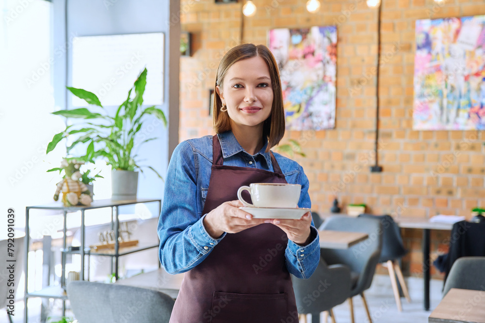 Young woman worker, owner in apron holding cup of coffee in restaurant, coffee shop Young woman worker, owner in apron holding cup of coffee in restaurant, coffee shop