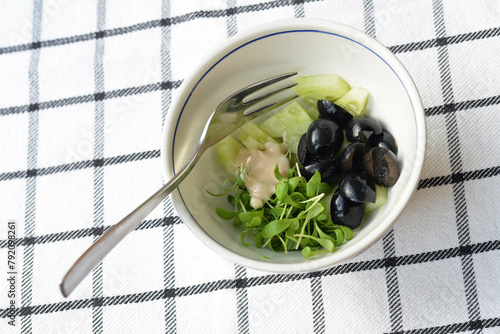 Green and black salad with cucumber, watercress and black olives with a drop of cocktail sauce and a fork in a white bowl on a black and white checkered cloth
