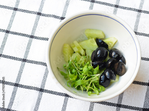 Green and black salad with cucumber, watercress and black olives in a white bowl on a black and white checkered cloth
