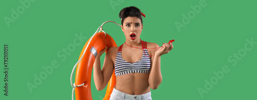 Shocked young pin-up lifeguard with ring buoy and whistle on green background