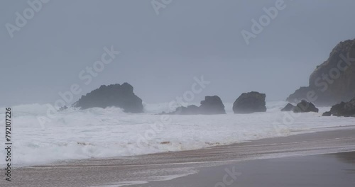 Cold winter ocean, sea beach with fogy extreme weather, huge waves, big rocks sticking out and maountains in the background. Dramatic gray landscape storm scene in Portugal. Splash wave in slow motion photo
